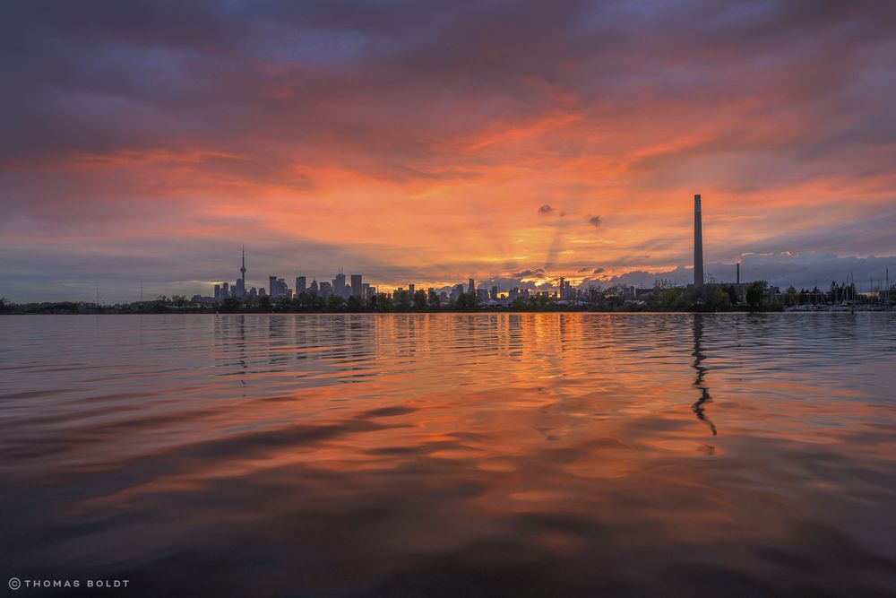 a photograph of a bright sunset over a gently rippling water surface that glows with reflected colour. the toronto skyline is visible on the horizon, including a tower of the ashbridges treatment plant, also visible as a rippled reflection. 