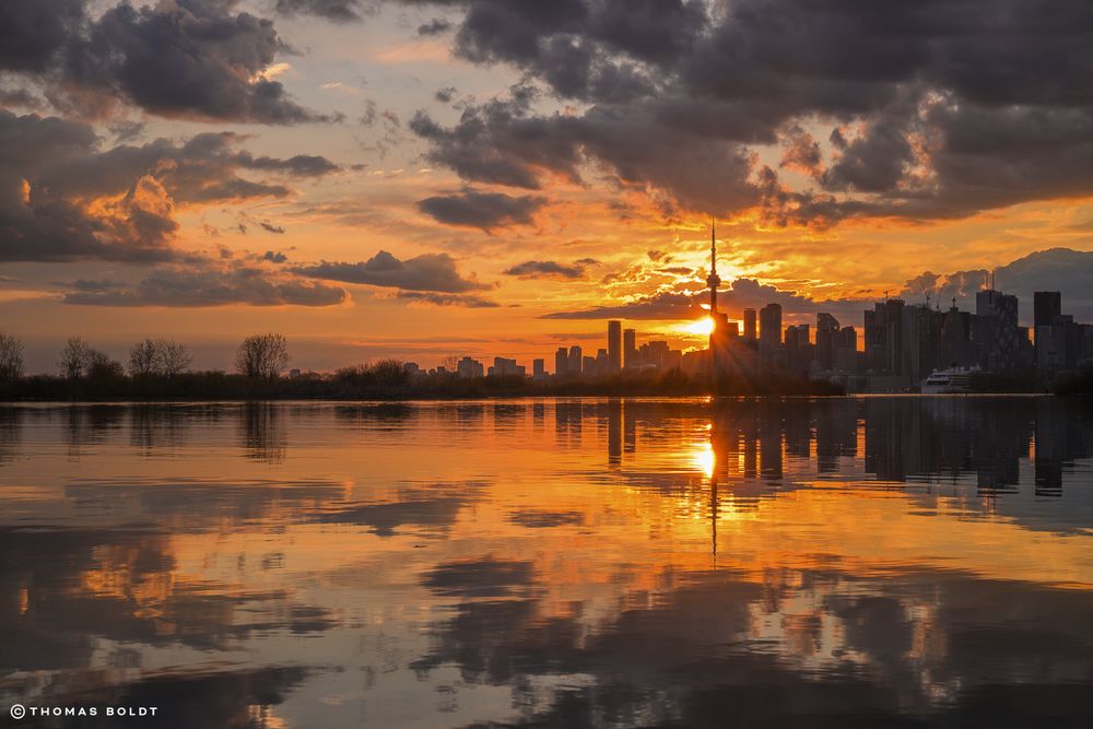 a photograph of the sunset on may 6, 2025 in toronto. the sun flares as it sets behind the CN tower, reflected in the still waters of toronto harbour 