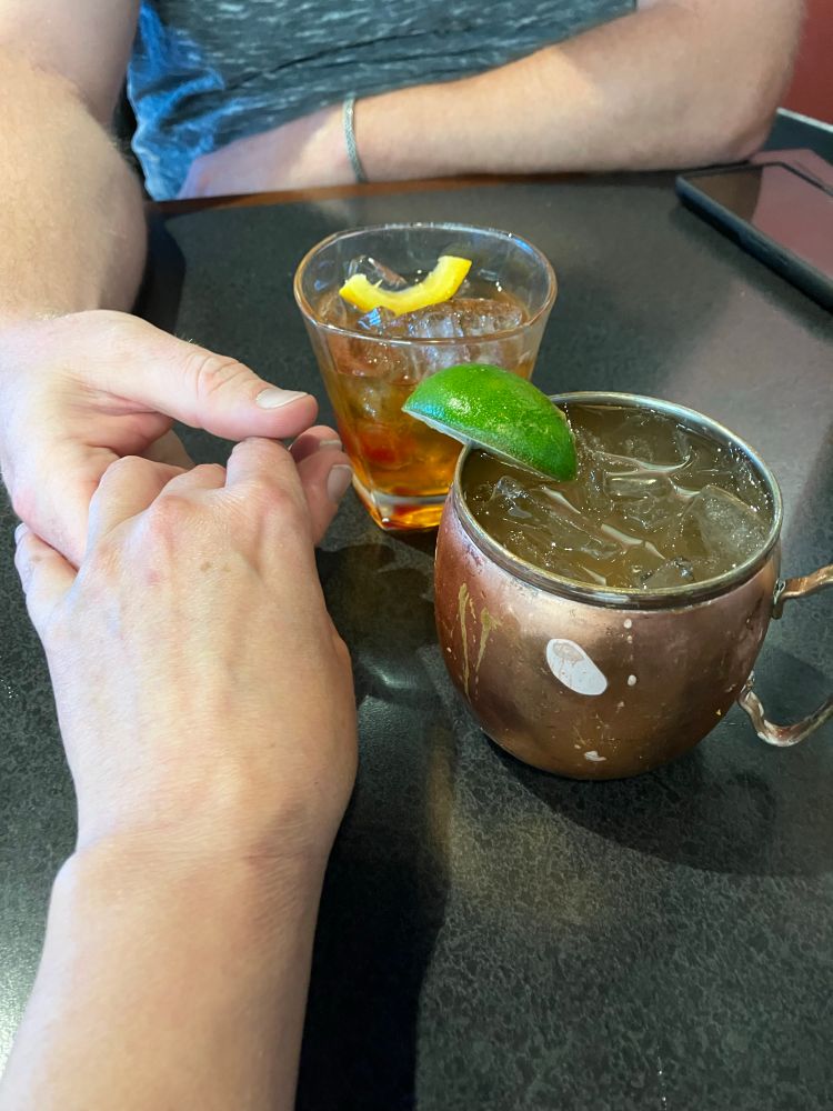 Two people holding hands in connection preparing for their flight. There is an Old Fashioned and a Bardenay Mule on the table next to their hands