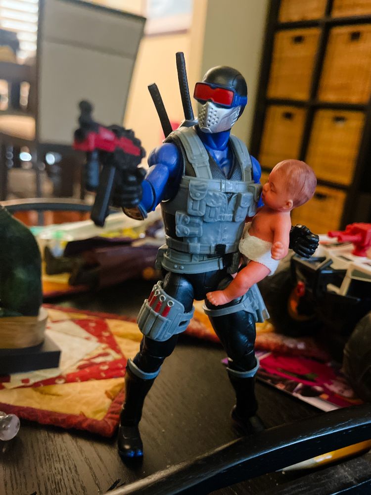 Snake Eyes in Blue costume with paintball mask and red goggles holds a large baby.