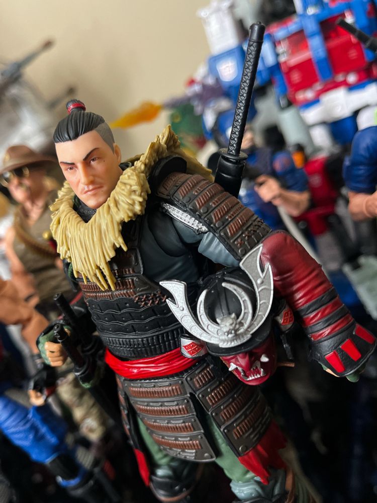 GI Joe Classified Budo with helmet off showing short haircut on the sides with the top in a knot. He’s got a grass cape.