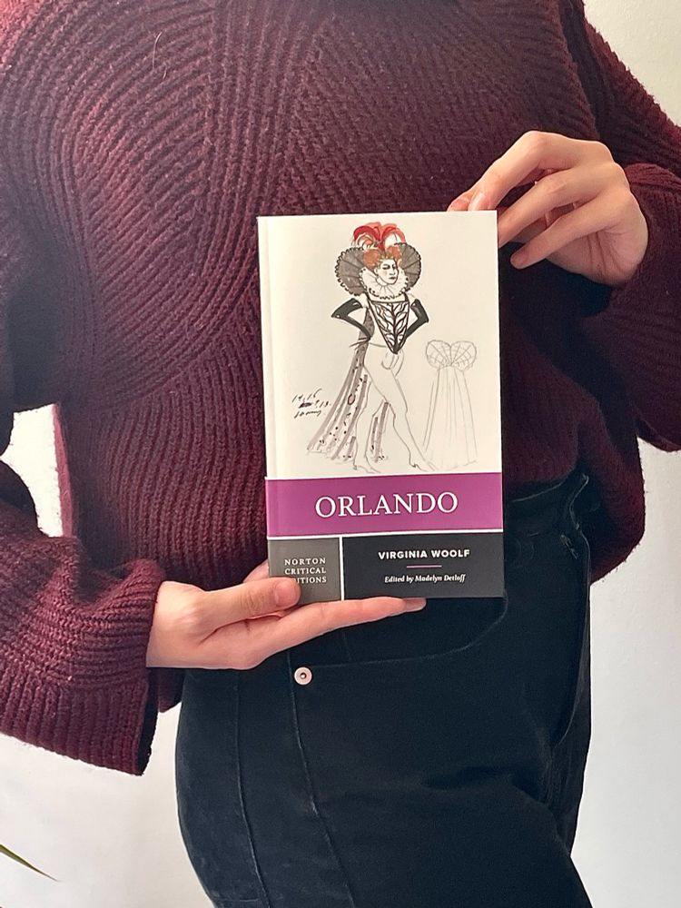 Woman in a purple sweater holding a copy of Orlando by Virginia Woolf. The book cover is a costume sketch from a stage adaptation of this work of queer LGBTQ fiction.