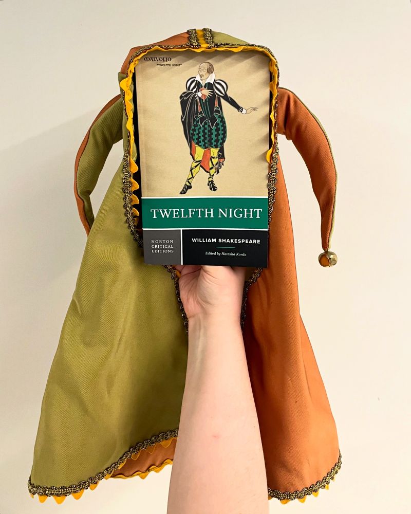 Twelfth Night: A Norton Critical Edition by William Shakespeare book wearing a green and orange jester hat. The cover image is of Dame Eileen Mayo’s costume design for Malcolm—a green outfit with yellow tights and a black cape