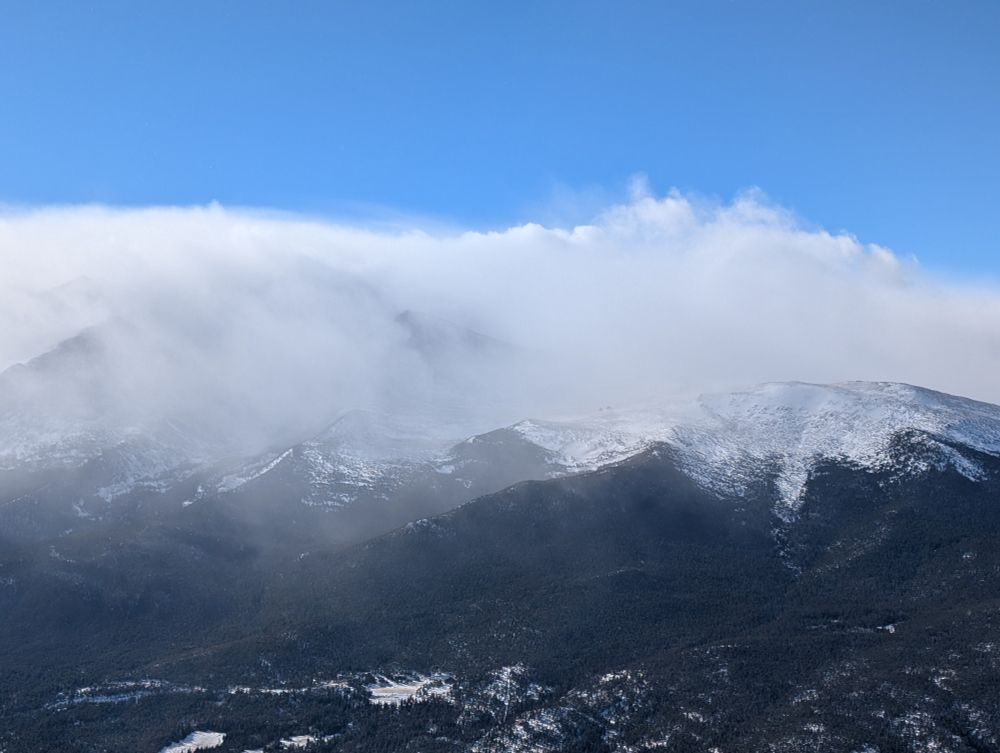 A series of snow-capped mountains (I think Lings Peak) obscured by cloud under a light blue sky