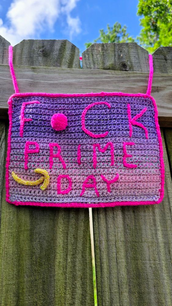 Handmade crochet craftivism hanging sign in purple, pink, and yellow that states F*ck Prime Day. Yarn: Spincycle and Lavender Lune. Icords created with Cordsmith by Size Inclusive collective.