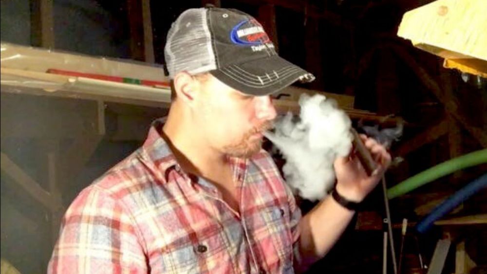 Craig Rauch in flannel shirt and trucker cap exhaling a thick cloud of smoke from his cigar