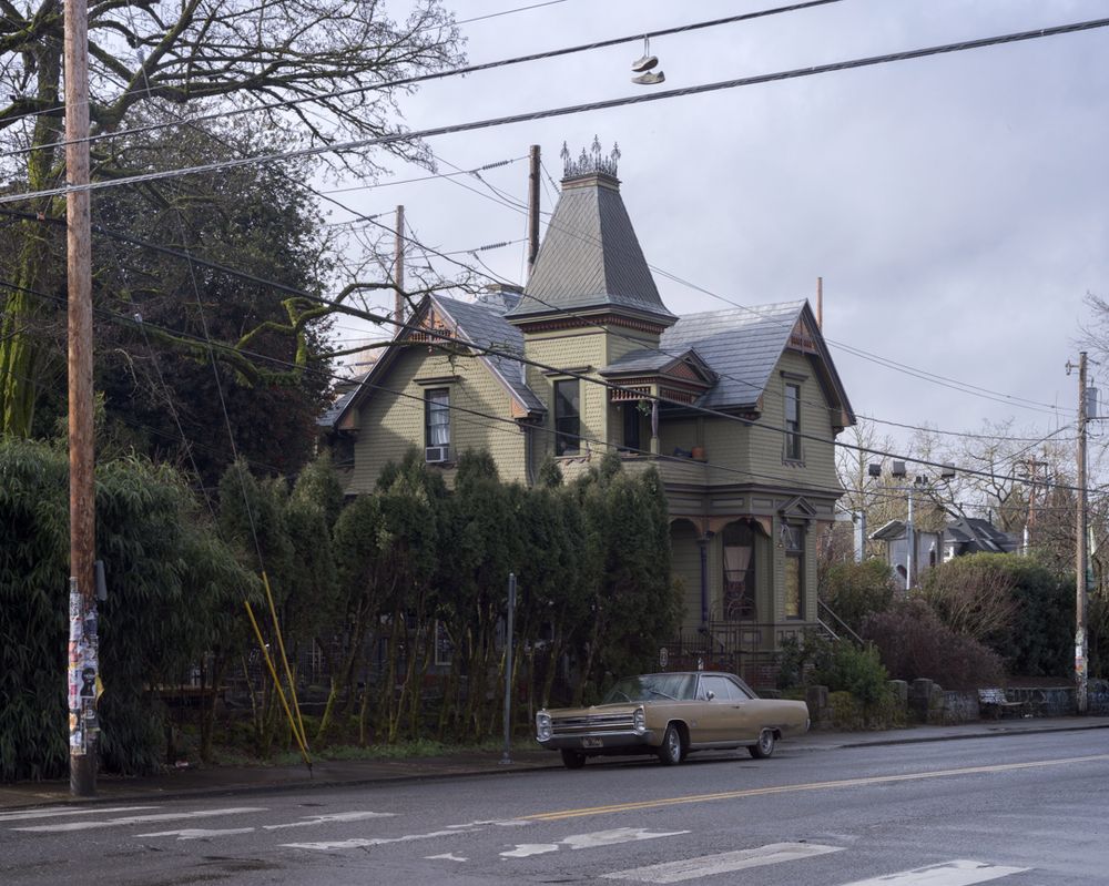 A dull green 1965-1968 Plymouth Sport Fury sits in front of a Victorian house painted a similar shade of green.  A pair of shoes hangs over the utility lines crossing the street in front of the house.