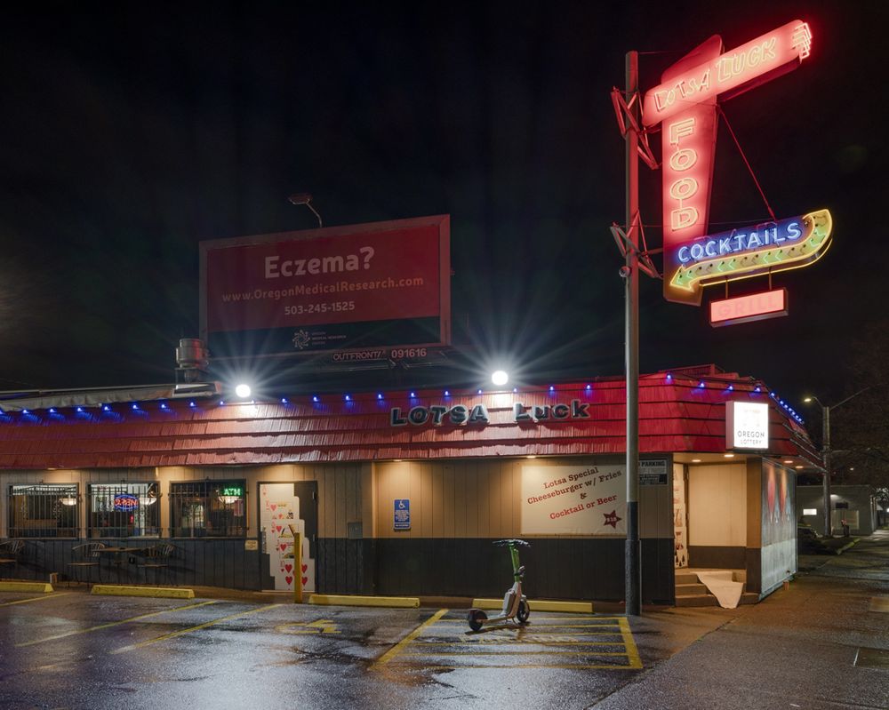 A bar called "Lotsa Luck," with a neon sign out front.  The door of the bar has images of a royal flush on it.  There's a Lime scooter parked in the disabled parking access aisle and an eczema billboard looms above.