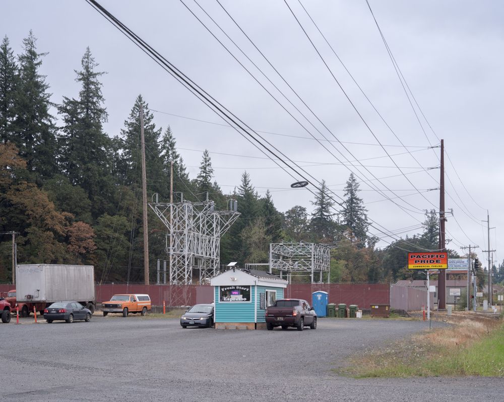 A drive-through coffee stand in the parking lot of a commercial fueling station has an electrical substation behind it. A line of trees is behind the the electrical substation.
