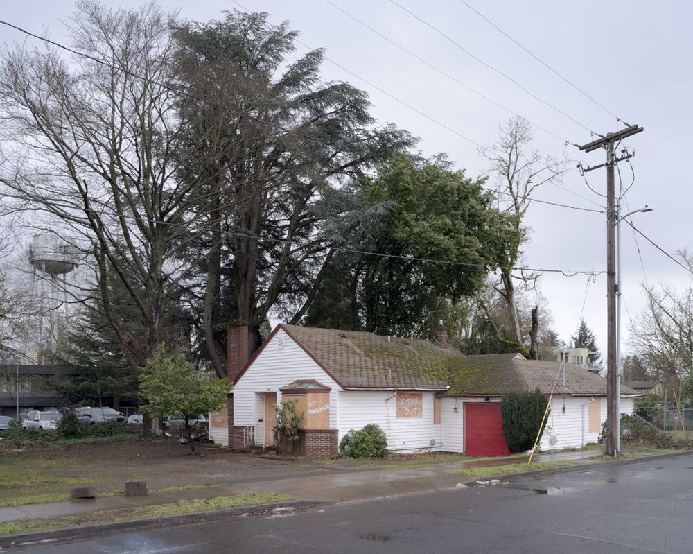 An abandoned house has boarded-up windows with "stay out" and "no trespassing" spray-painted on the boards. There is a utility pole in front of the house, trees behind the house, and a water tower in the middle distance behind the trees.