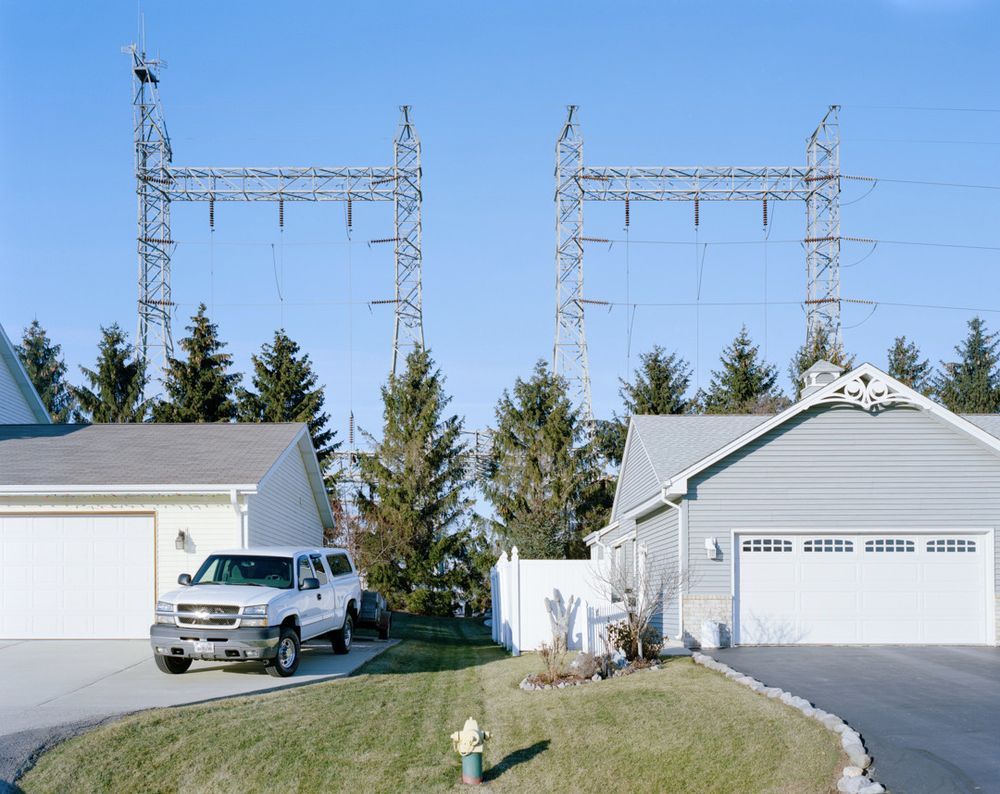 A well-manicured lawn with a fire hydrant on it separates two new-build houses.  Behind the houses is a line of trees, and behind the line of trees are two large high-voltage transmission towers connecting to a substation underneath.