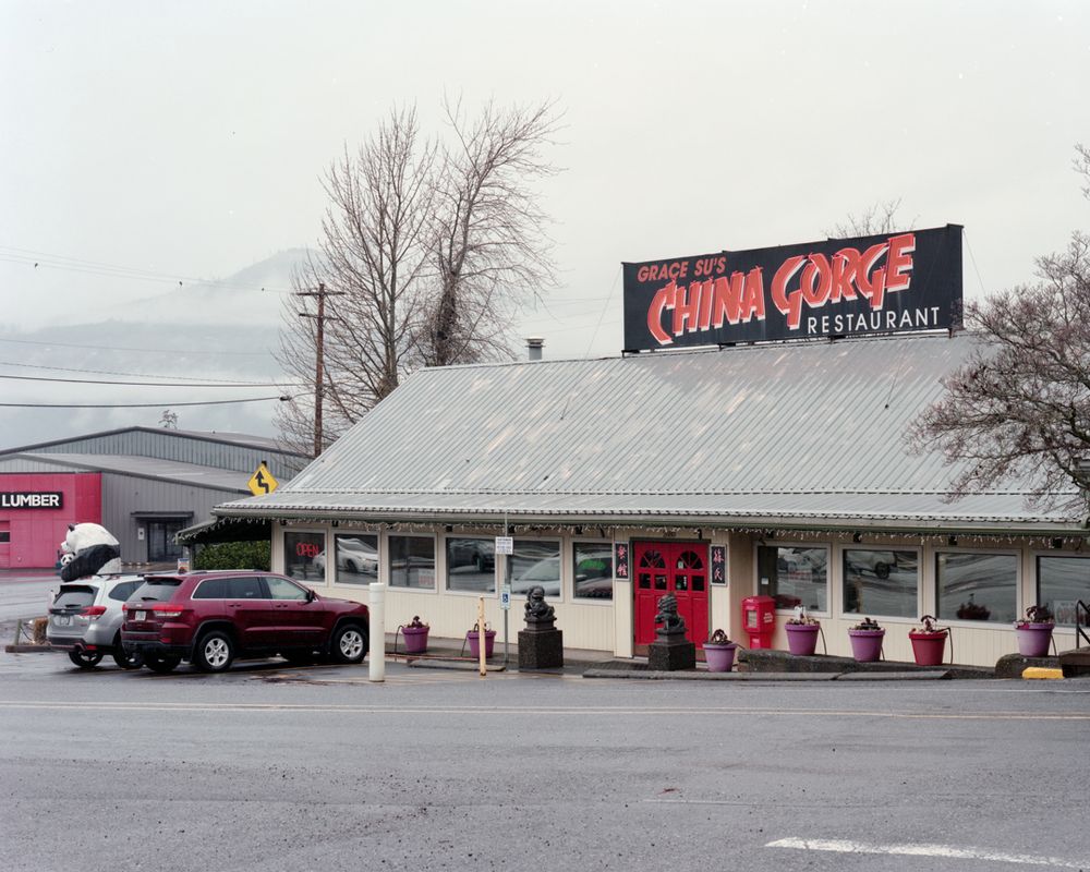 A Chinese restaurant with a large sign on a cloudy day. There is a lumber store across the street and a mountain peeks out of the clouds in the background.