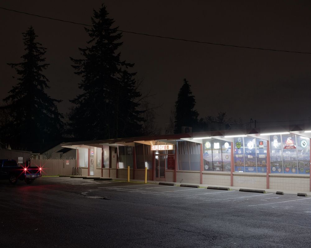 a Portland strip mall at night. evergreens in the background pierce the dim city light reflecting off the bottom of the clouds