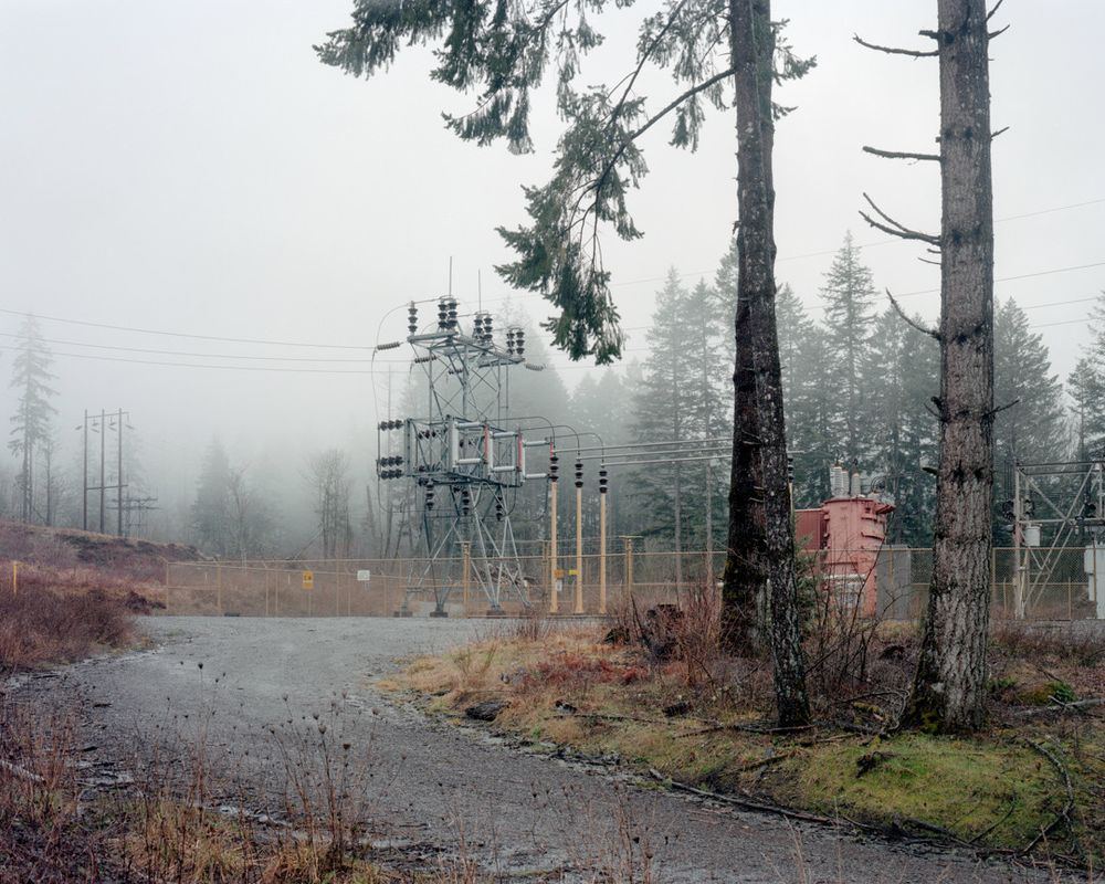 An electrical substation at the end of a gravel road is surrounded by trees and shrouded in mist.