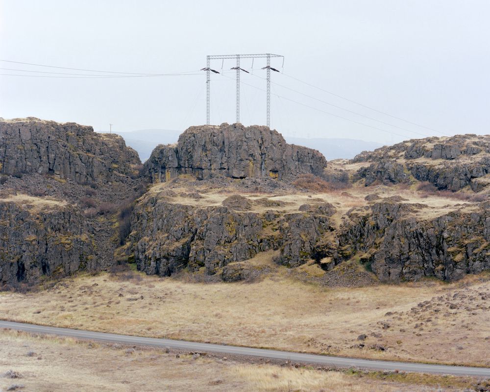A high-voltage electrical transmission tower stands on a rocky outcropping surrounded by dry grass on a cloudy day.