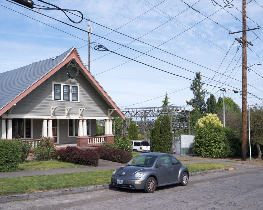A Portland Craftsman bungalow has a substation and a cell tower in the lot next to it. There's a grey VW New Beetle parked on the street in front.