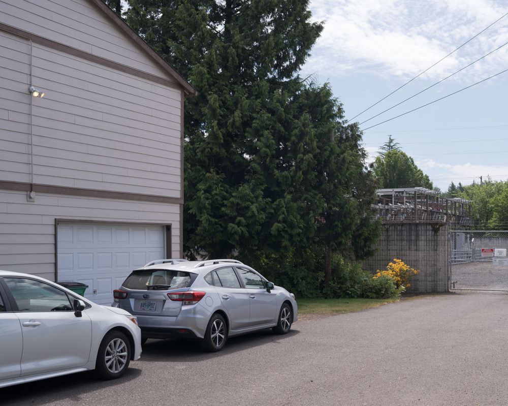 Two cars are parked next to a residential building. The road the cars are on leads to the gate of an electrical substation, which is partly hidden by trees behind the residential building.
