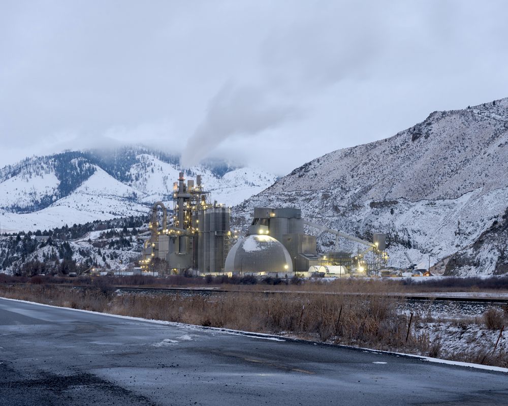 A cement plant emits steam at twilight, nestled beside snowy mountains.