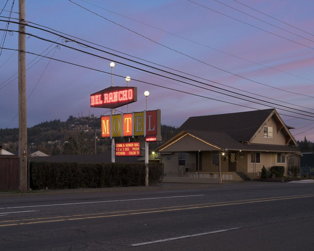 The Del Rancho Motel has a 50s-style mostly-working sign with neon. It's on a wide street, and in the background there's a mountain with houses and radio towers on it. The sky is pastel with the last light of blue hour.