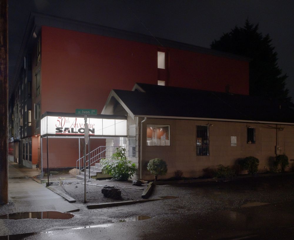 A hair salon in a house with a brightly lit canopy over the entrance stands in front of a new-build apartment building at night.