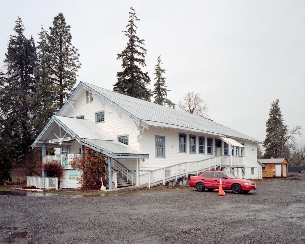 A white building with a "Rockford Grange" sign in front of trees. A bright red early 90s Subaru Legacy sedan is parked next to orange traffic cones in the gravel lot that surrounds the front of the building.