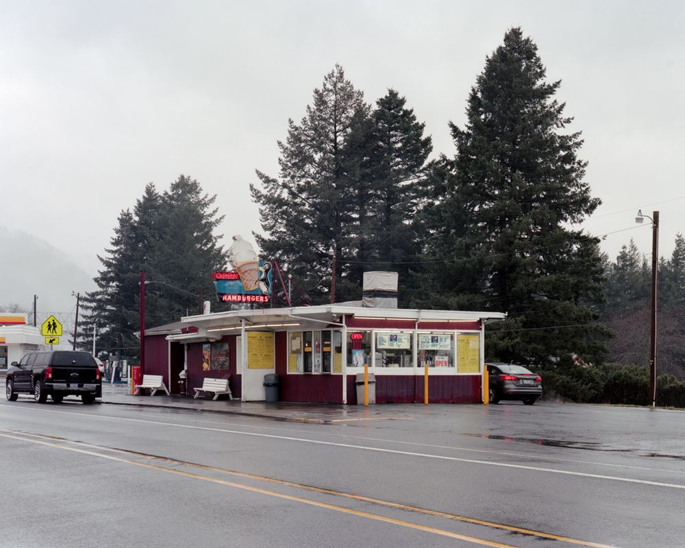 A drive-through hamburger restaurant with a neon sign depicting a penguin carrying a soft-serve ice cream cone.  Evergreen trees stand in a row behind it, and behind those are a bridge and a hillside shrouded by cloud.