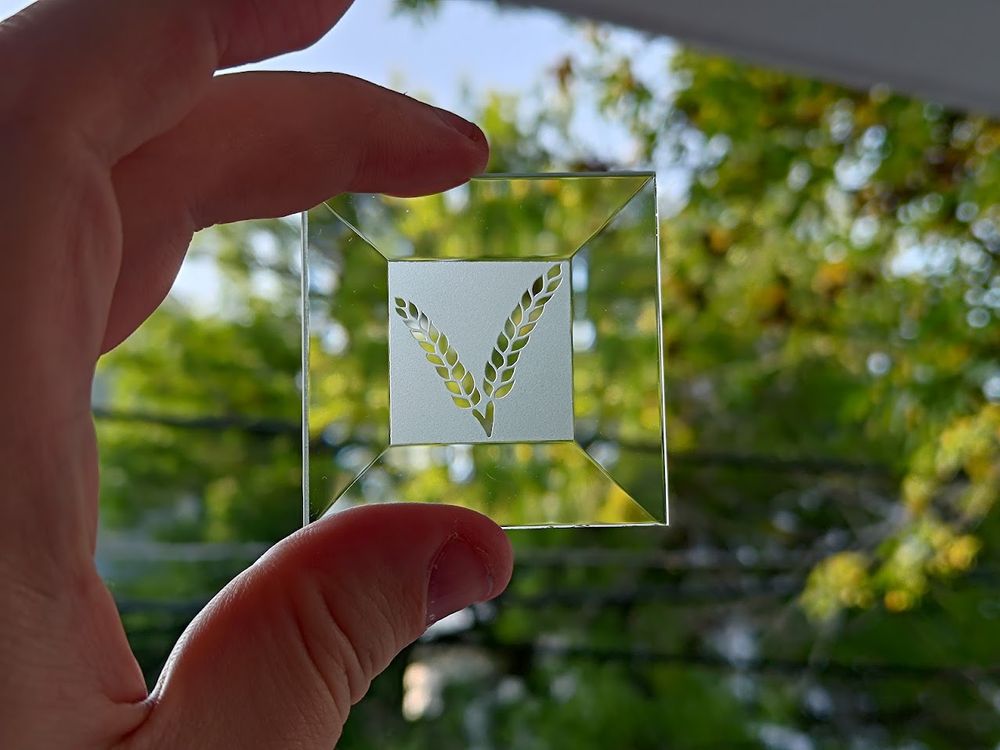 A square glass bevel. The center portion has been sand-etched so that the majority of it is frosted. The shape of two pieces of wheat are left clear.