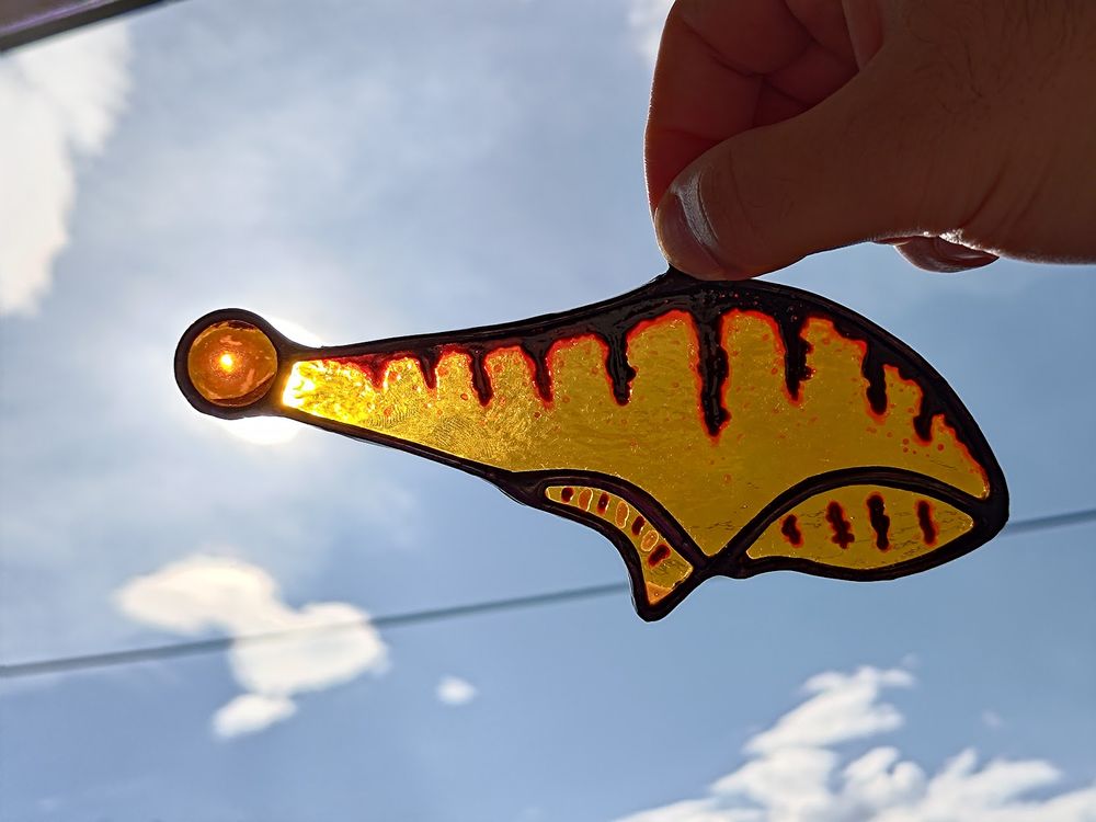 A stained glass version of a Ytram (frog) from the game Riven. Orange water glass with paints to show the marking from the in-game animal. It has a round orange gem for an eye/head. It is held against a sunny sky, with the sun coming through the gem.