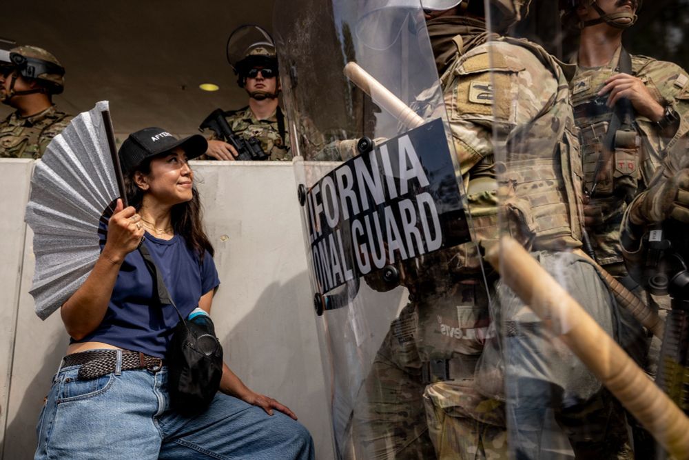 Photo from the San Francisco Chronicle showing a No Kings Day protester facing off with national guard. The national guard are heavy armed and behind riot shields. The protester is a young woman, absolutely unbothered, holding a large fan up by her shoulder and eyeing the NG with a look of bemusement.