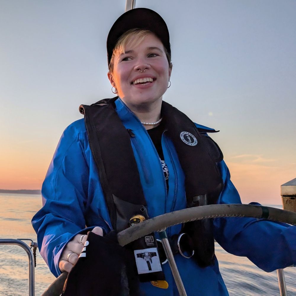 A white lesbian in a blue windbreaker behind the wheel of a boat.