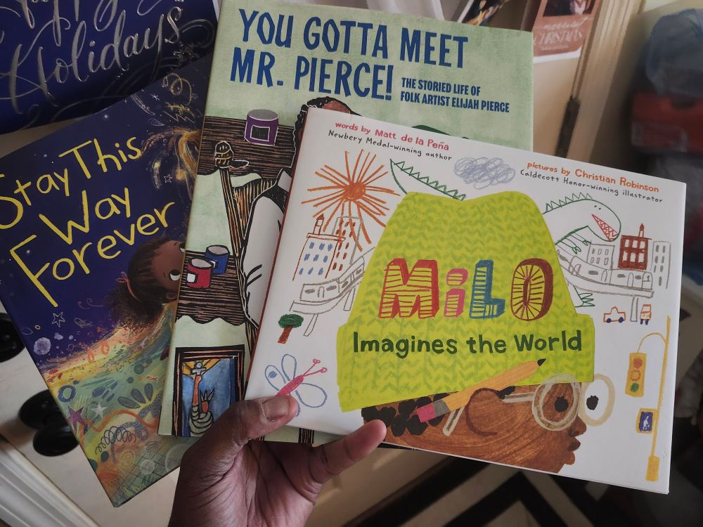 A photo of a brown hand holding 3 children's books in front of a white door with holiday cards on the door. The book titles are Milo Imagines the World, You Gotta Meet Mr. Pierce!, and Stay This Way Forever.