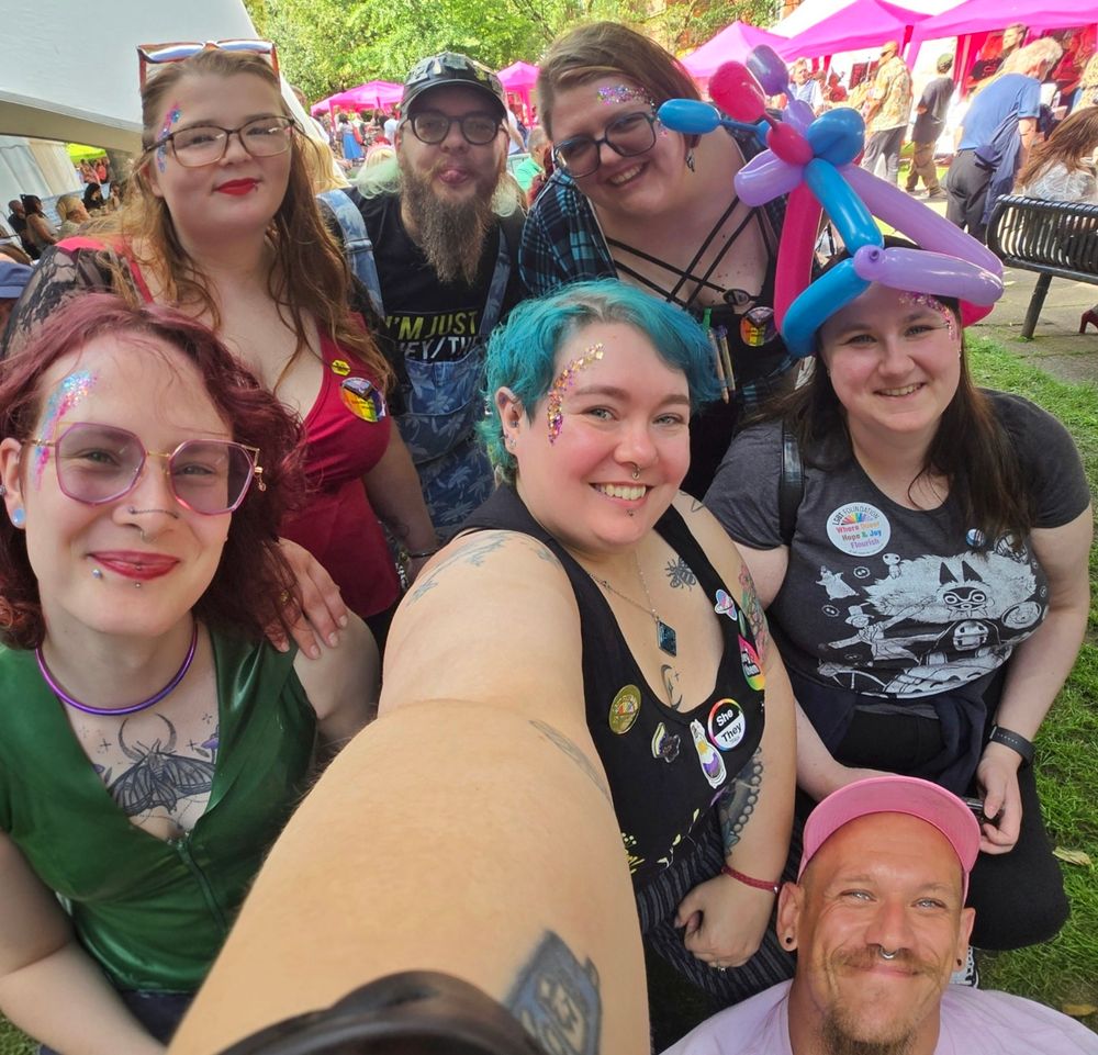 A group selfie of 7 people, all happy looking into the camera. Several of them have glitter on their faces, and once is wearing a balloon crown. 
