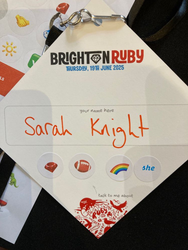 Brighton Ruby lanyard with hand written name “Sarah Knight” and stickers of emojis in the “talk to me about” section. Emojis are: Ruby, American Football, Rainbow and the word “she” in blue.