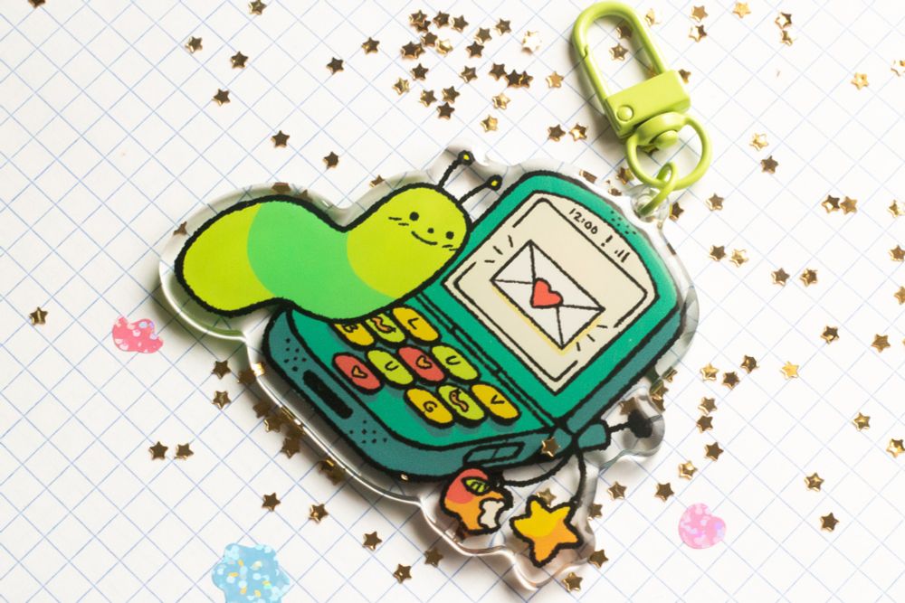 an angled picture of the luv bug keychain against a white grid background, surrounded by smaller glittery stickers and gold star glitter