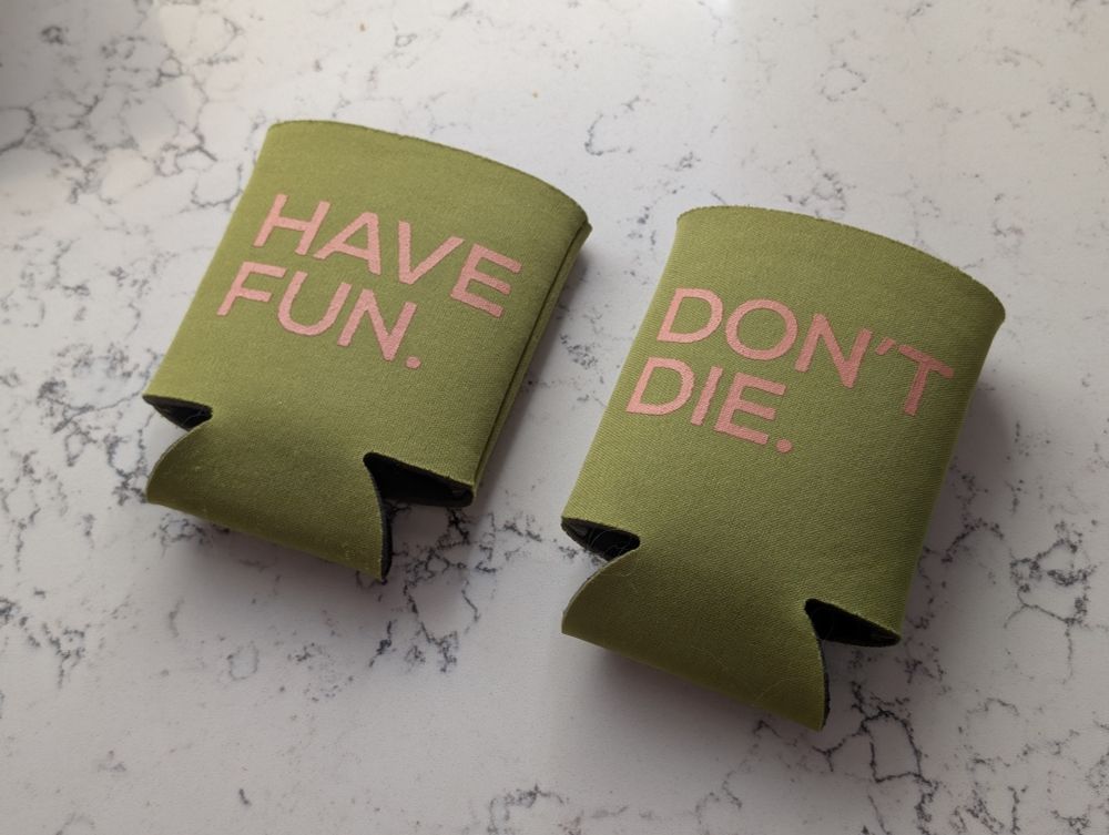 Two drink koozies, both olive green with a peachy pink font. One says (in all caps) Have fun. The other reads Don't die.
