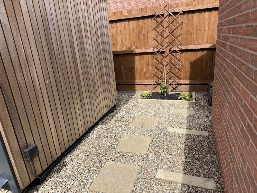 Photo showing a sandy coloured gravel area with stepping stones towards a small raised border with new plants and trellis. It’s all between a wood side of a shed on the left and a garage wall on the right 