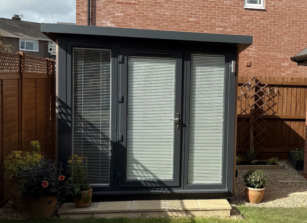 Photo of the front of an anthracite coloured garden office with a trench door. To the left of the step are three potted plants of different types and sizes and there is a smaller potted plant to the right, all sat on sandy coloured gravel