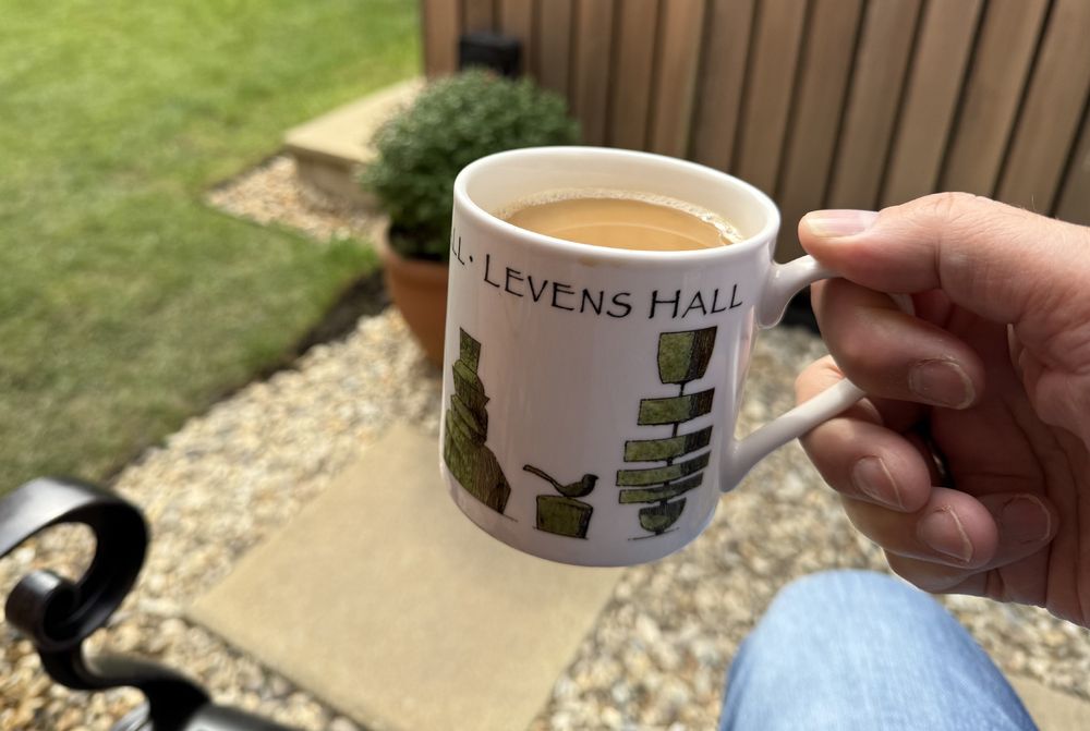 Photo of a hand holding a mug of coffee in a Levens Hall mug in front of some gravel and a plant. Behind is some red cedar cladding