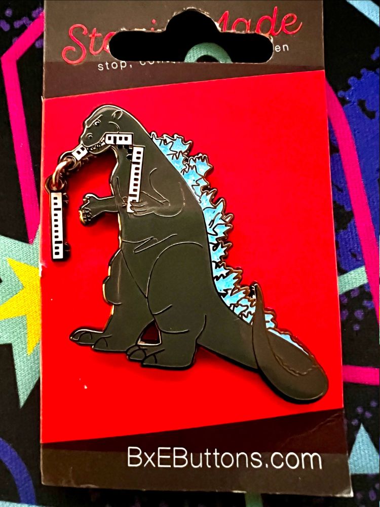 An enamel pin of Godzilla with a train in his maw, with a train car dangling free on a small chain length. The printed URL on the backing paper BxEButtons.com leads to the maker's etsy page.