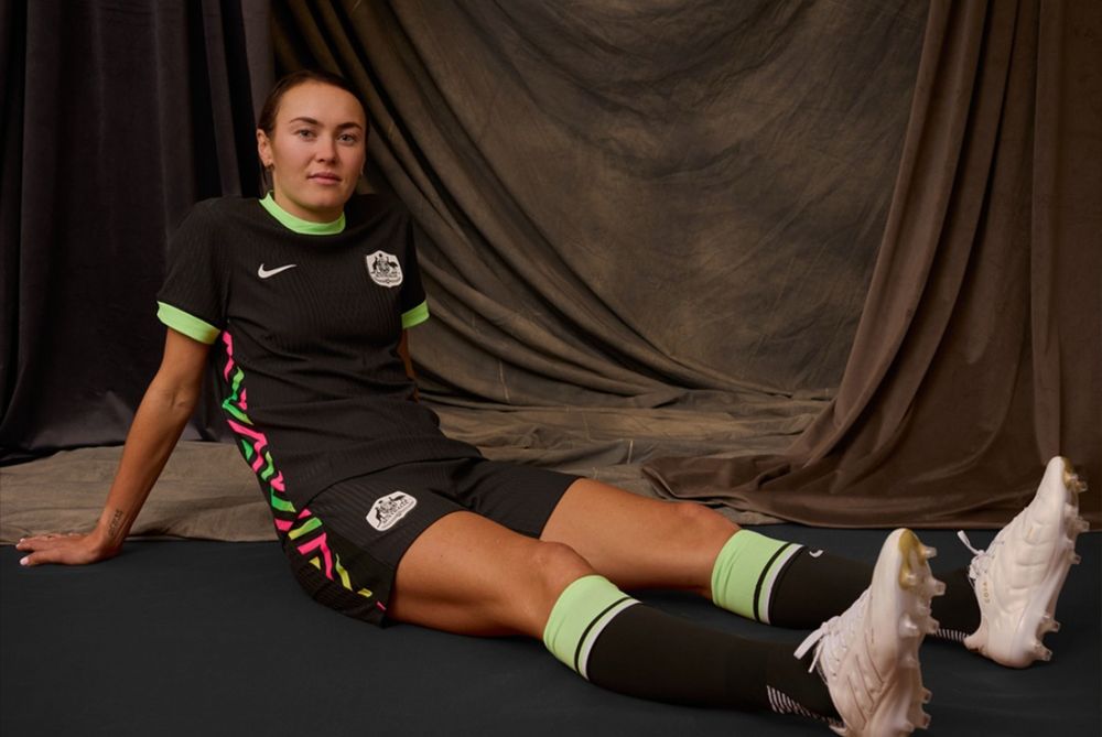 Australian forward Caitlin Foord is sitting with her legs out in front of her, leaning back on her hands. She’s wearing a new away kit in black with neon accents. She looks 💯 