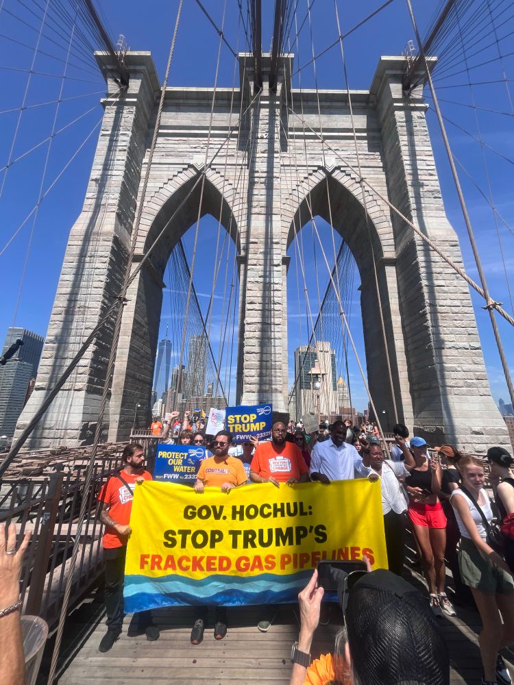 Gov Hochul: stop trumps fracked gas pipelines says banner with people crowded behind it on the walkway of the Brooklyn bridge with the bridge arches in the background.