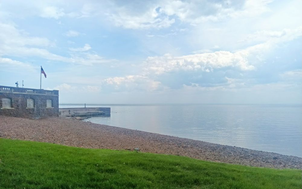 A picture of the shoreline of Lake Superior at the historic (haha by American standards at least) Glensheen Mansion. The stone boathouse is to the left and the glassy water is reflecting fluffy white clouds. The beach is made of small pebbles and stones.