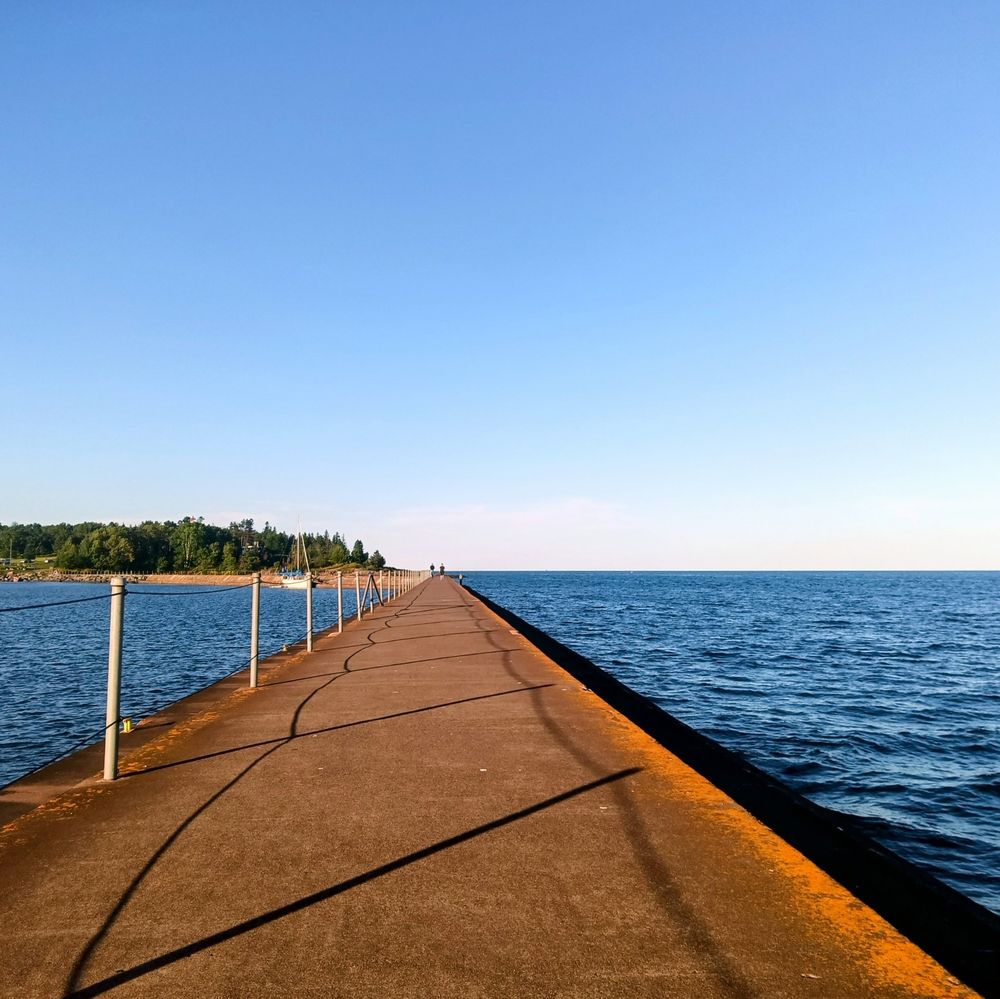 The cement breakwall / pier in Two Harbors. There is bright blue water on either side of the pier and pine trees off to the shore on the left and you can't see the shoreline at all on the right!