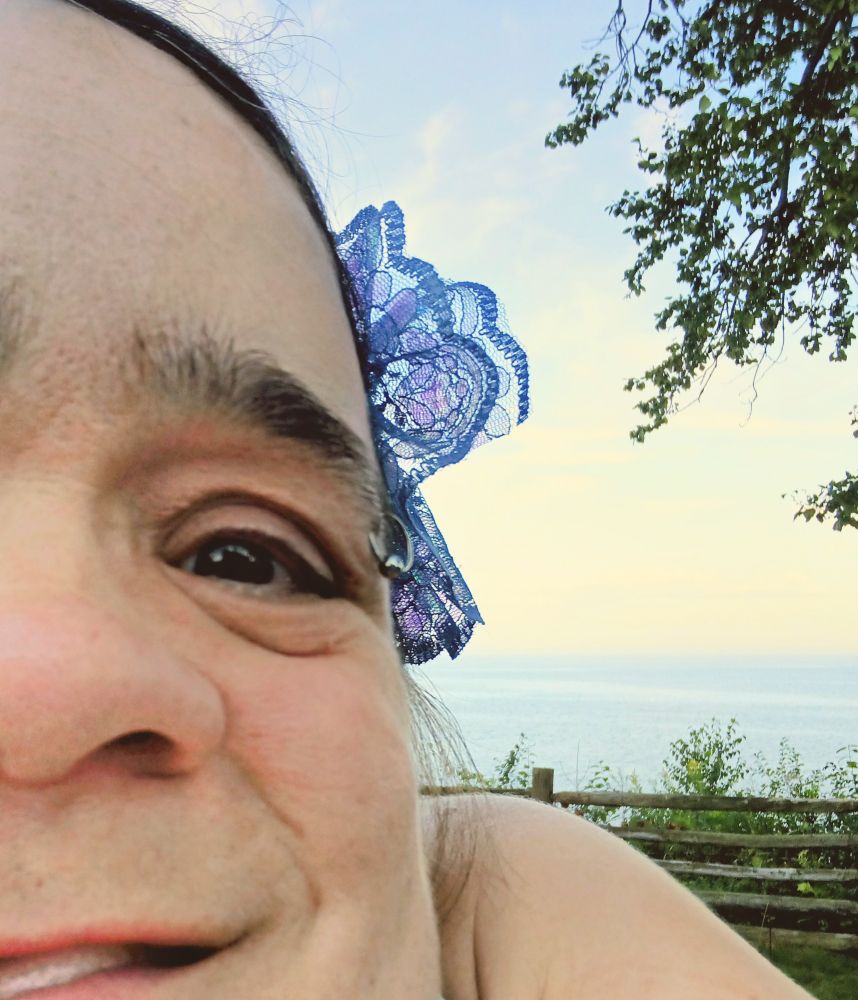 Gaelynn taking a selfie by the lake on their apartment patio. She has a blue flower clip in her hair and you can only see one eye in the photo because her arms are short and she can't hold the phone very far away haha