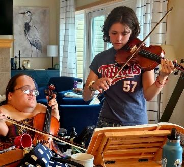 Gaelynn playing fiddle with her niece Naomi. Gaelynn has her violin upright in her wheelchair, like a tiny cello, and her niece is holding her violin the regular way. They are both reading the sheet music for a traditional tune in The Fiddlers Fake Book called Give Me Your Hand.