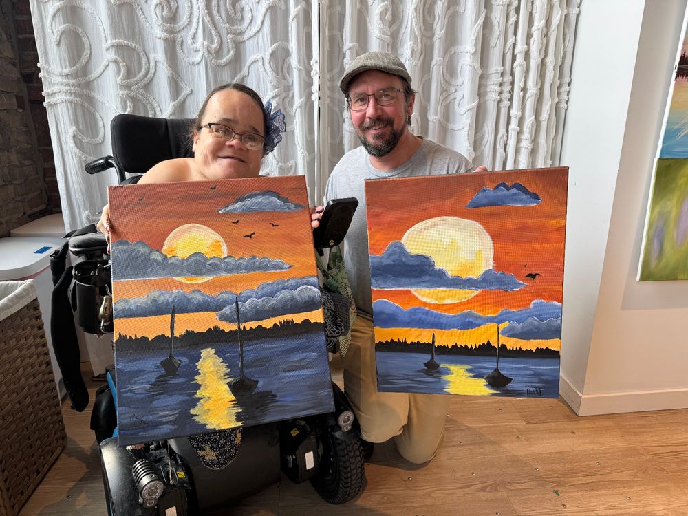 Paul and Gaelynn pose side by side holding 18x24 inch canvases and smiling. Each painting shows two sailboats on the lake with the sun setting and clouds in the sky. Paul's sun is bigger than Gaelynn's (he is a Leo, after all). 🌅
