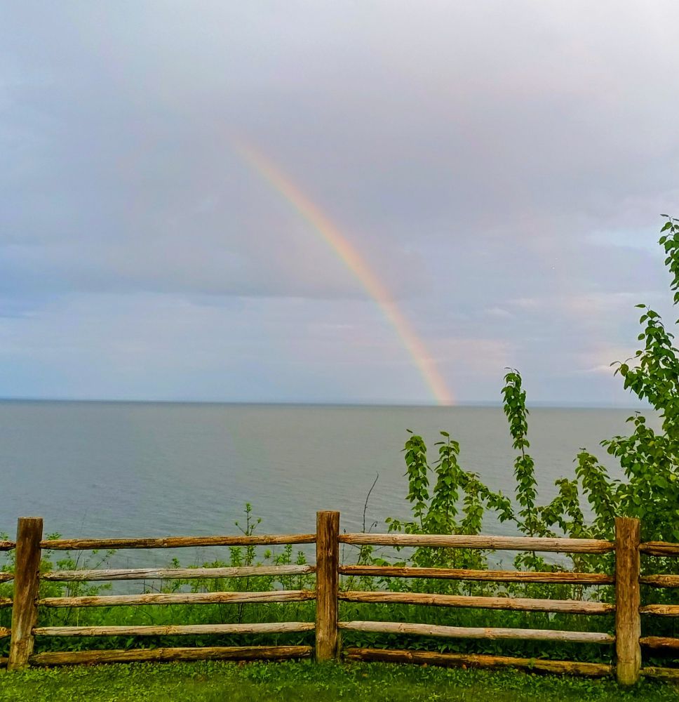 A rainbow over the calm waters of Lake Superior. In the foreground is a wooden fence and some bushes