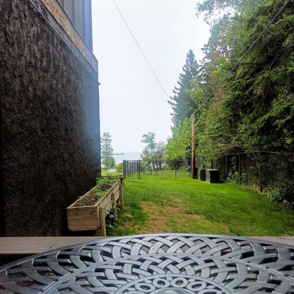 A view from Gaelynn's tiny patio. You can see the edge of her metal patio table and grass and trees in the distance. Way out there is Lake Superior, you can see an ore boat passing by, partially hidden by a pine tree. 