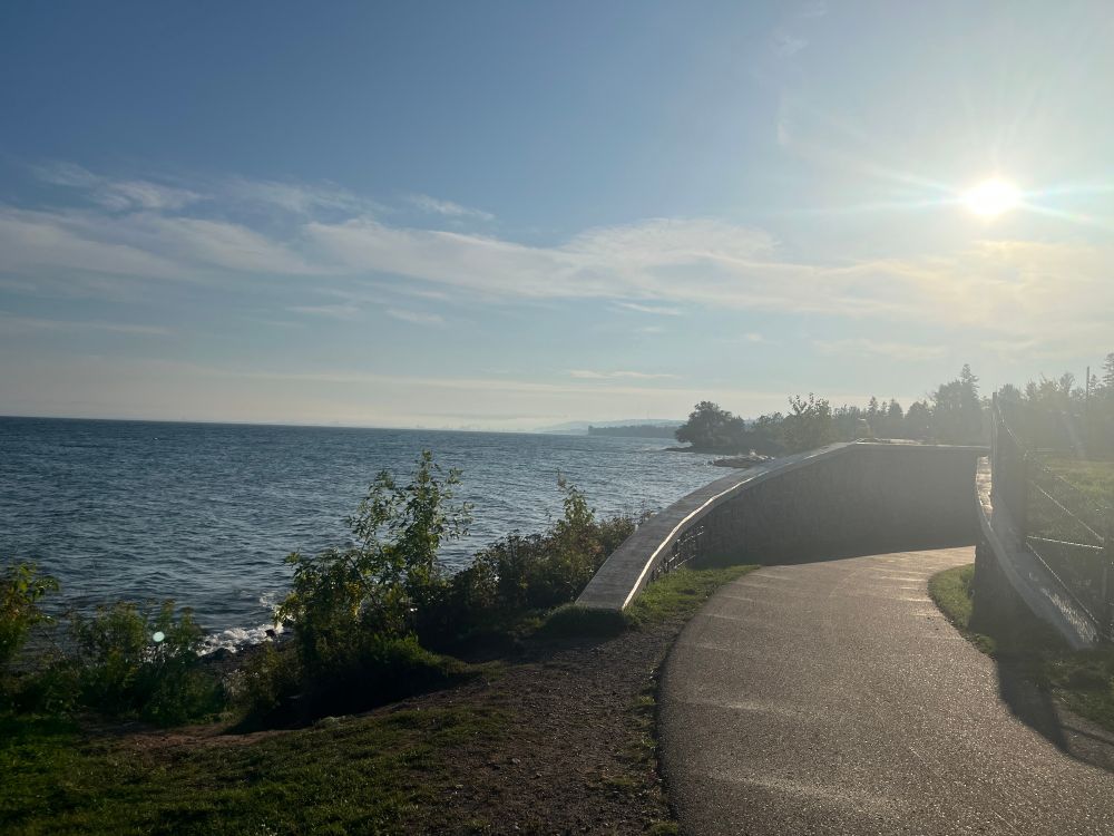A picture of the shoreline of Lake Superior with the sun shining high on the right hand side. You can see a tiny outline of the Arial Lift Bridge in the distance if you look close enough, shrouded in mist.