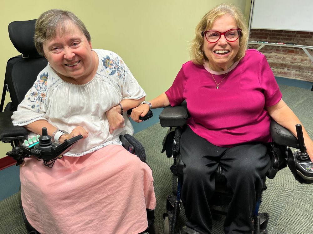 ID: In front of a yellow wall, Carrie and Judy sit side-by-side in their electric wheelchairs. Judy is wearing black pants and a fuchsia shirt and Carrie is wearing a pink skirt and a white top with green and pink embroidery on the shoulders. Judy has her hand on Carrie's armrest. Both are smiling.